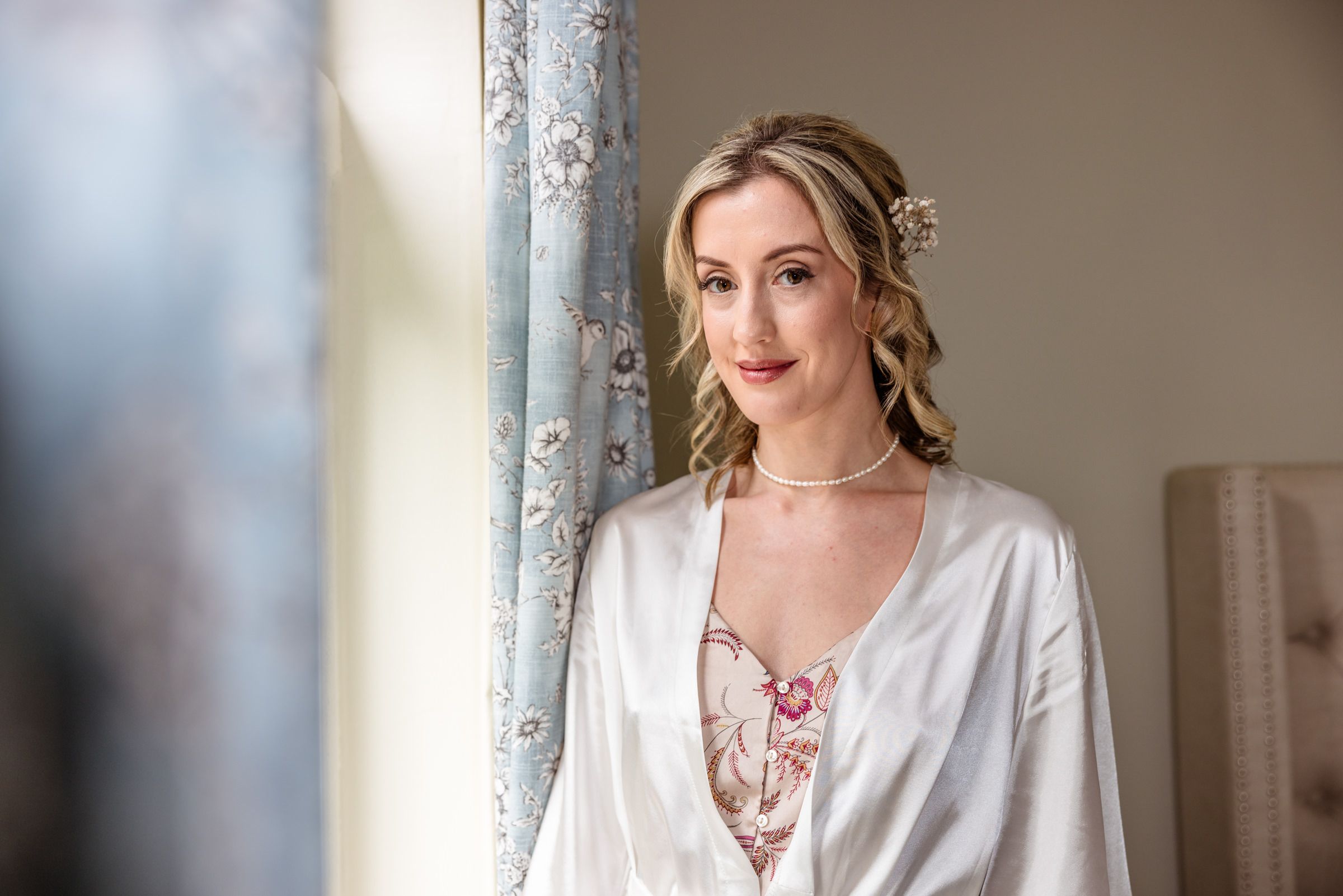 A bride in a white silk robe poses elegantly by a window during wedding preparations.
