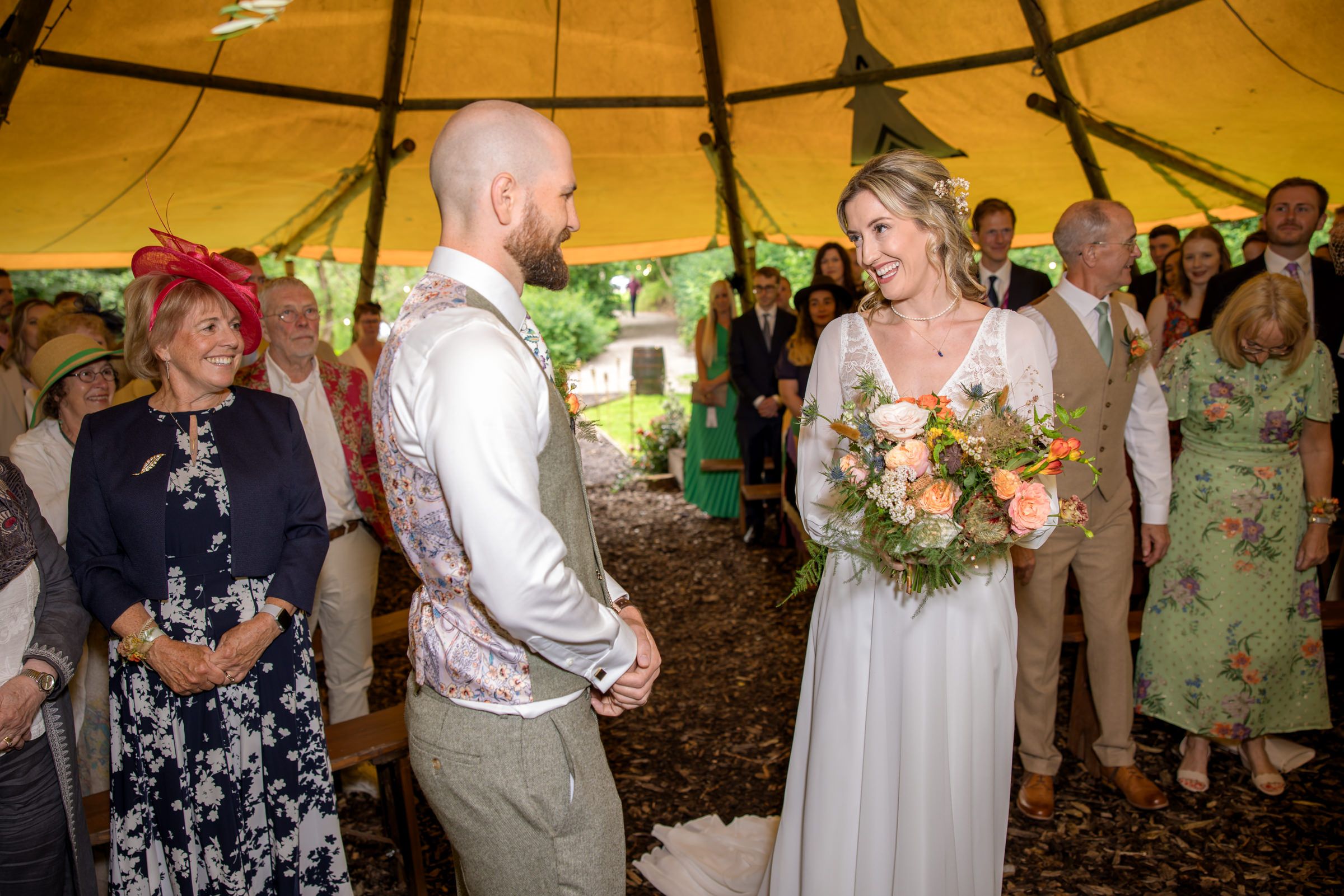 Wedding ceremony in progress under a yellow canvas tent with guests watching.