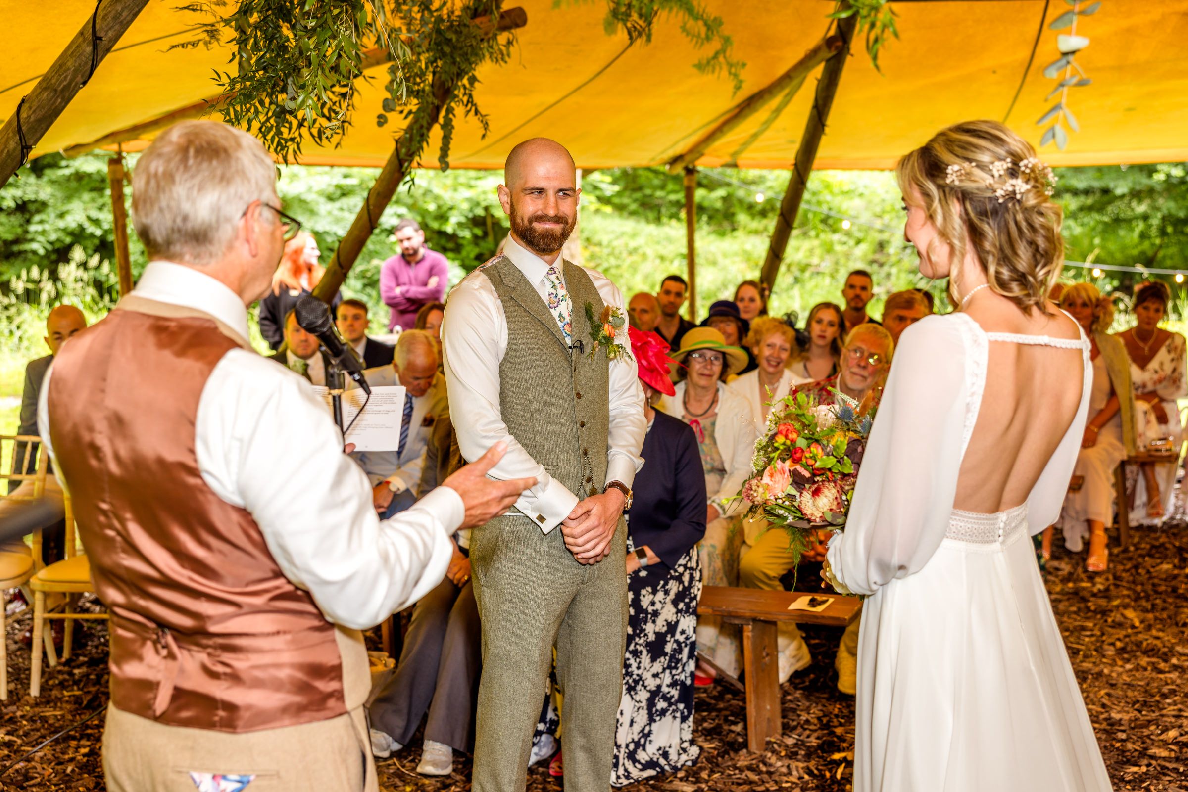 A wedding ceremony takes place under a yellow canopy tent with guests seated in a woodland setting.