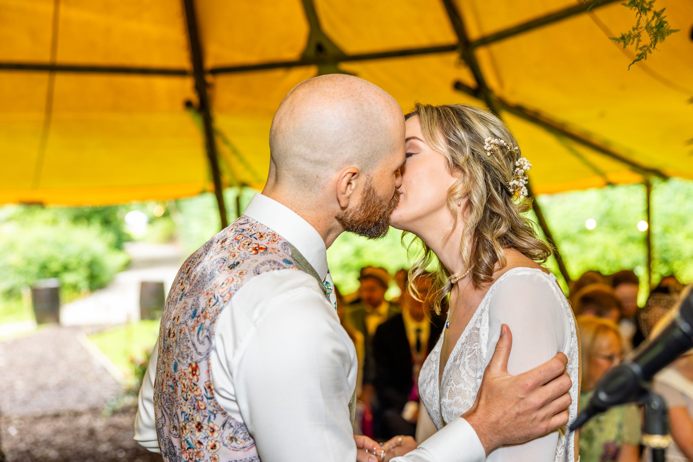 A couple shares their first kiss under a vibrant yellow tent canopy during an outdoor wedding ceremony.