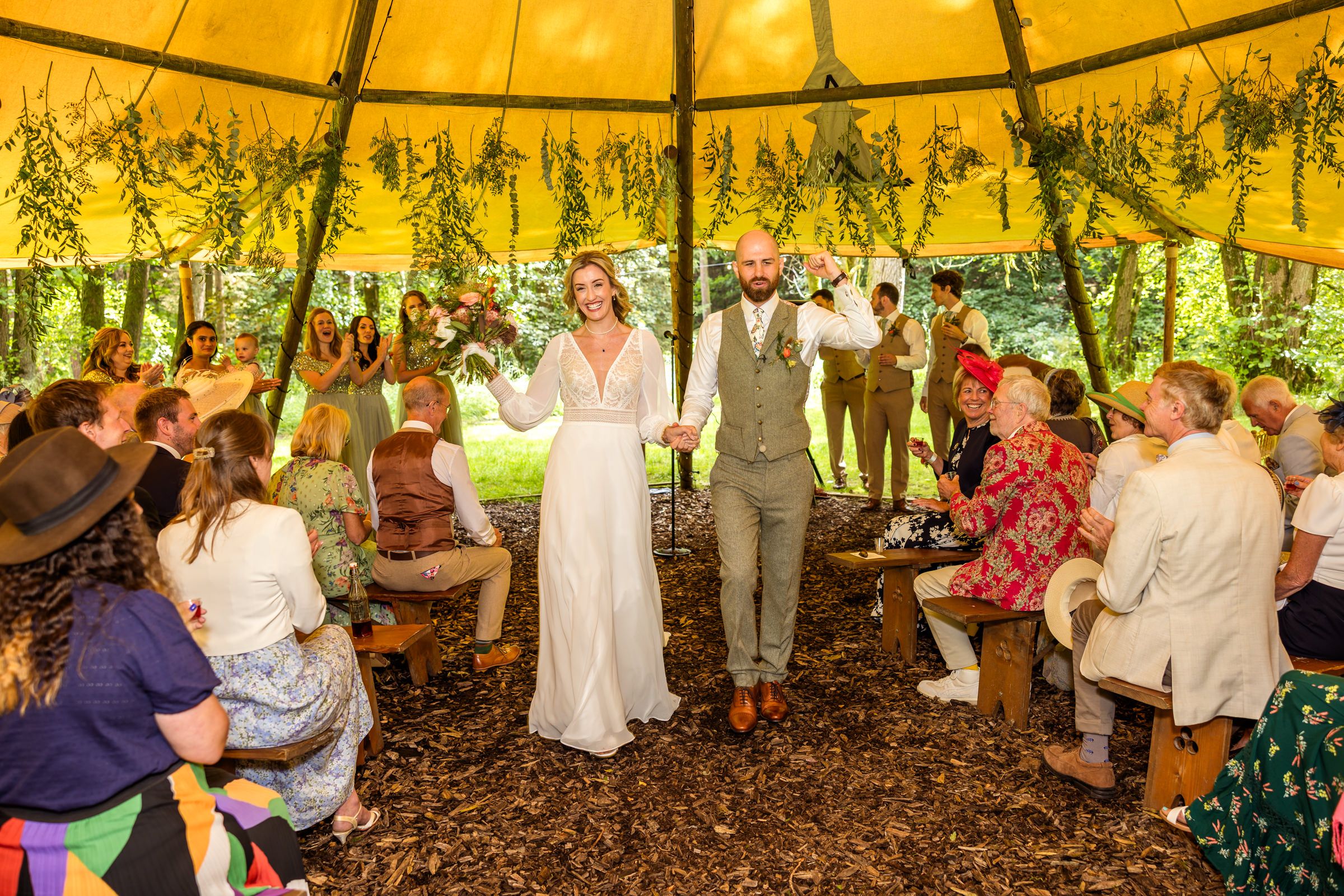Wedding celebration under a yellow tent with hanging greenery as guests applaud the newlyweds.