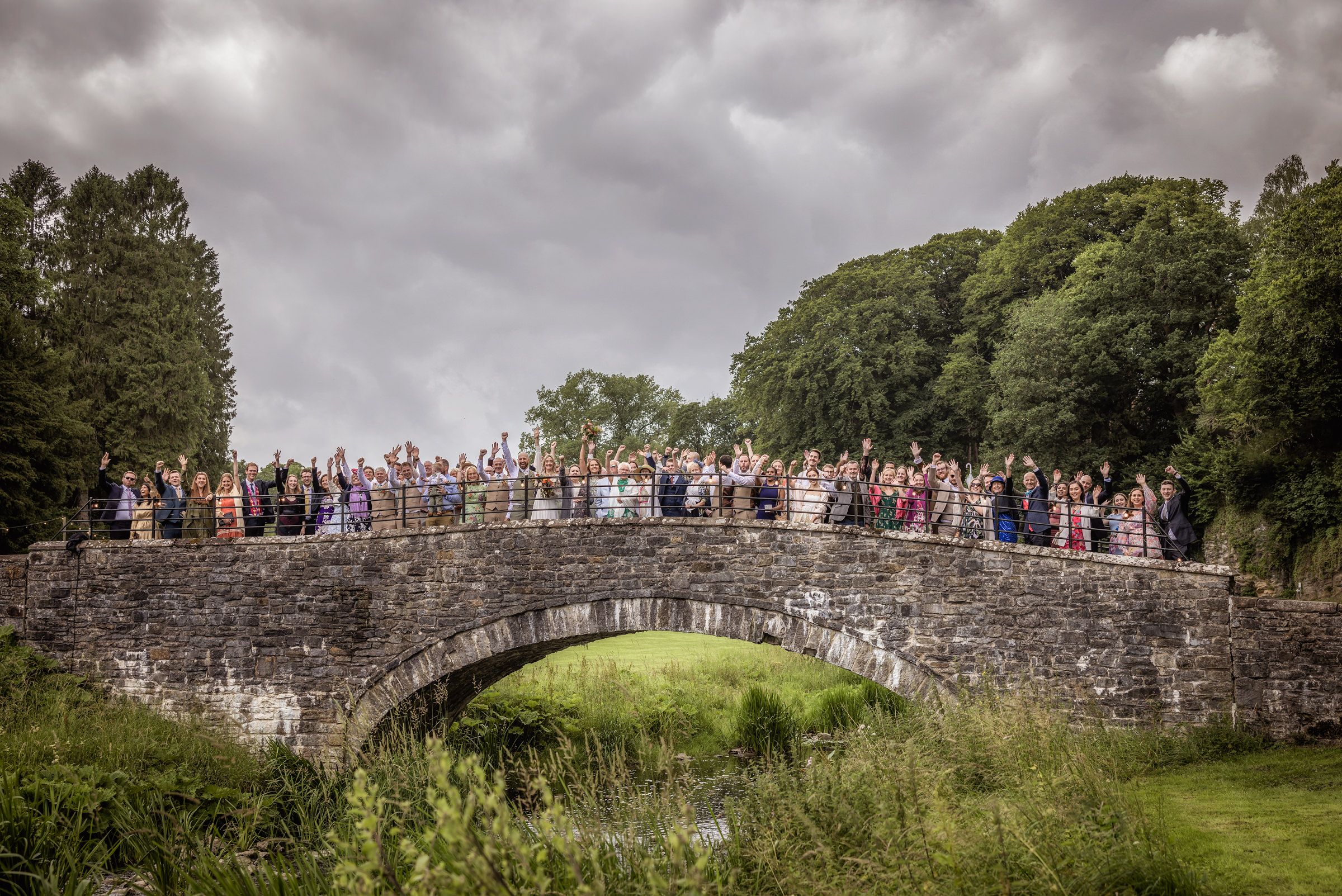 Large wedding group gathered on a rustic stone bridge in scenic countryside setting.