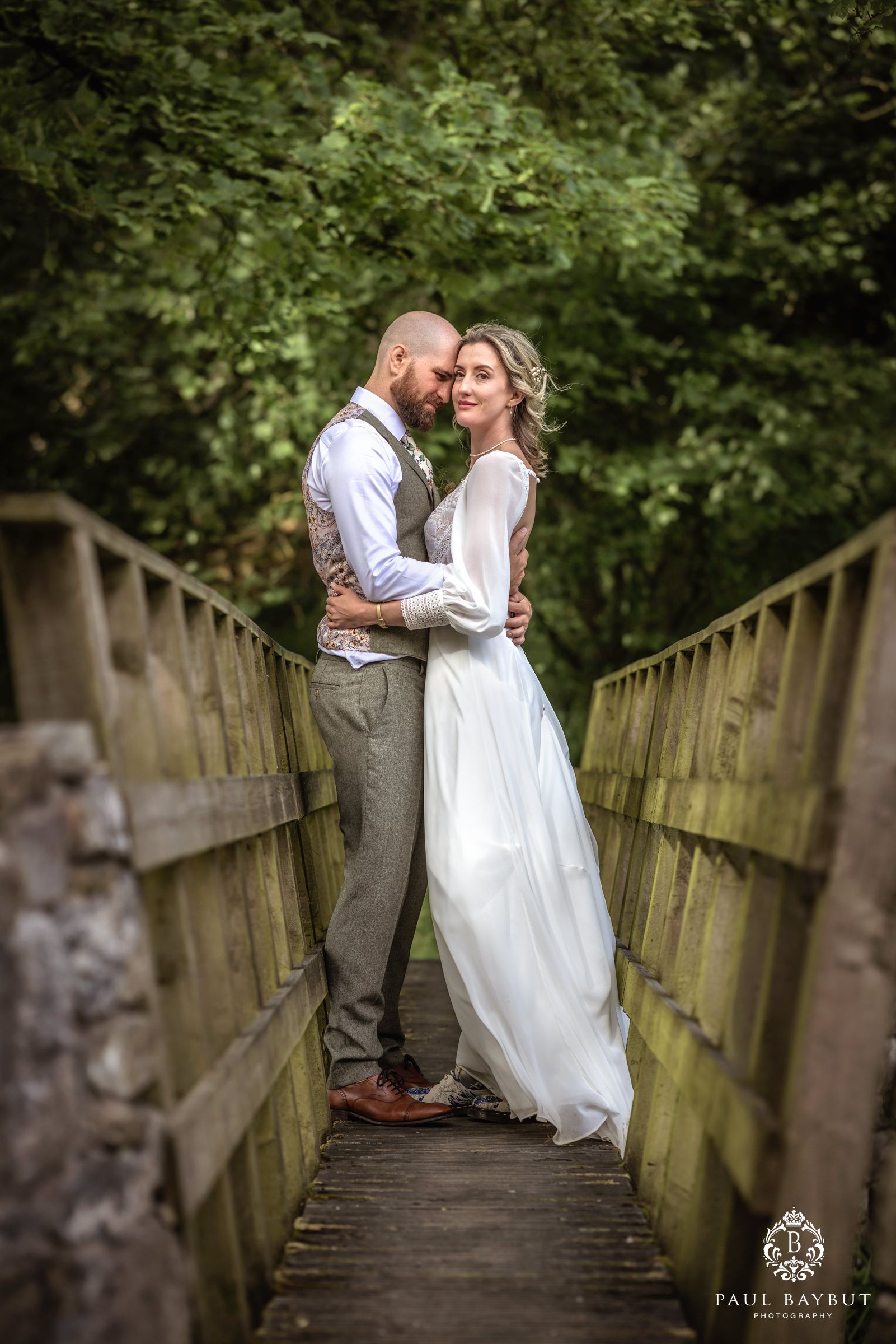Romantic wedding portrait on wooden footbridge surrounded by lush greenery.