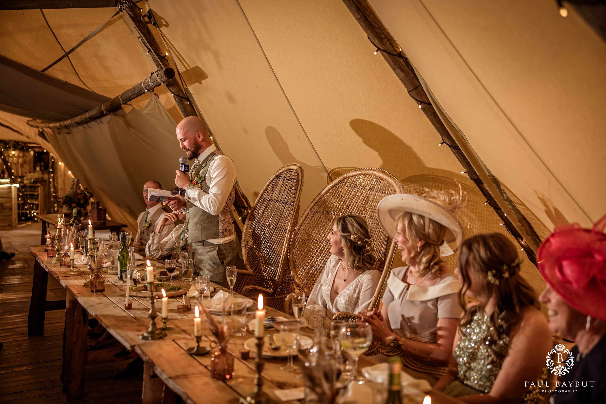 Wedding guests seated at a long table under a canvas tent with vintage-style decor and candlelight.