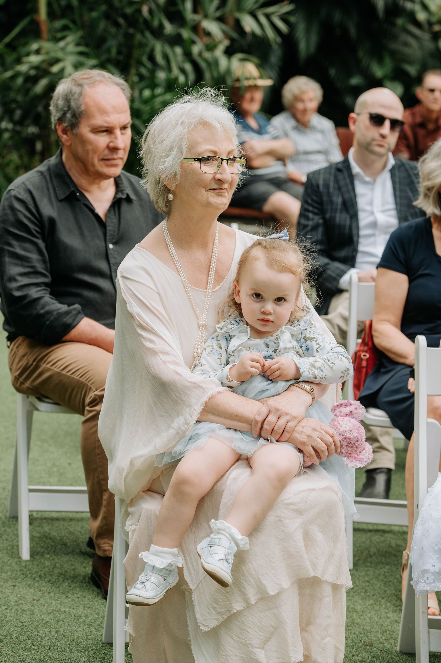 Wedding guests seated on white chairs enjoying outdoor garden ceremony.
