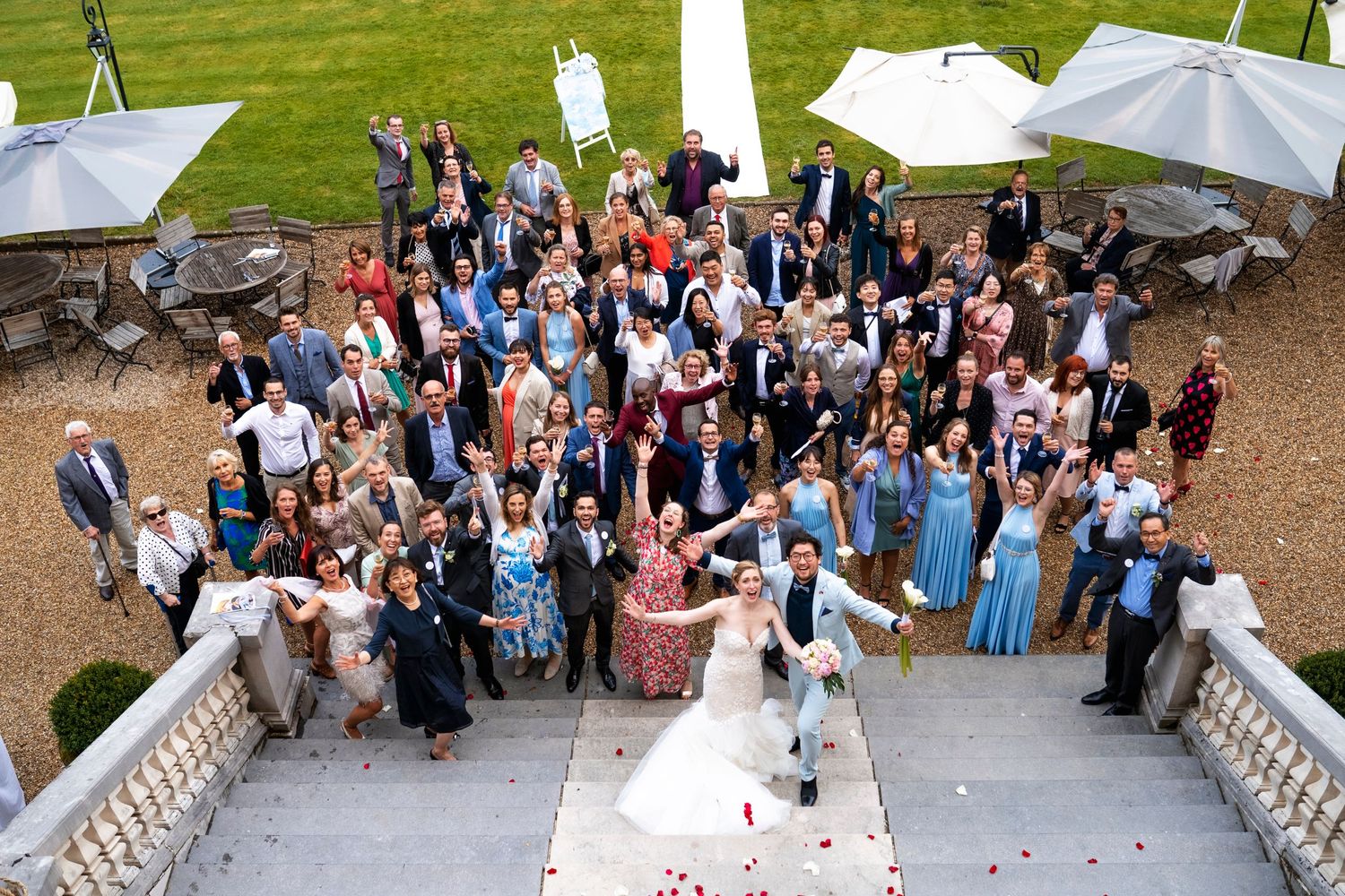 A large wedding group photo captured from above on elegant stone steps with white tent canopies.