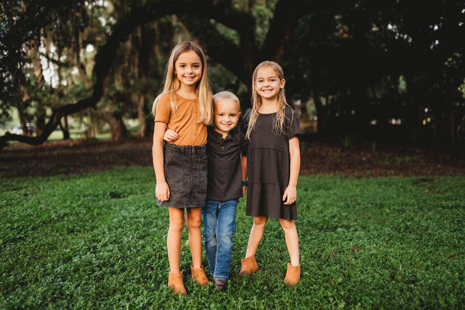 Three children standing together on grass wearing coordinated fall outfits in brown and orange tones.