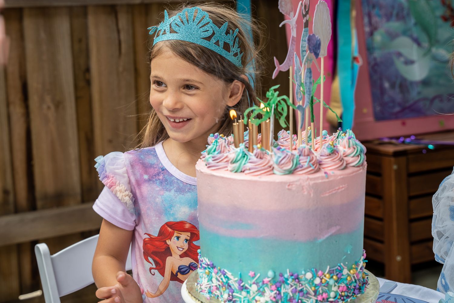 Happy young girl celebrating birthday with large mermaid-themed pink and blue cake.
