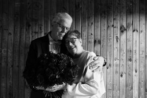 A couple embraces while holding flowers against wooden paneling in black and white.