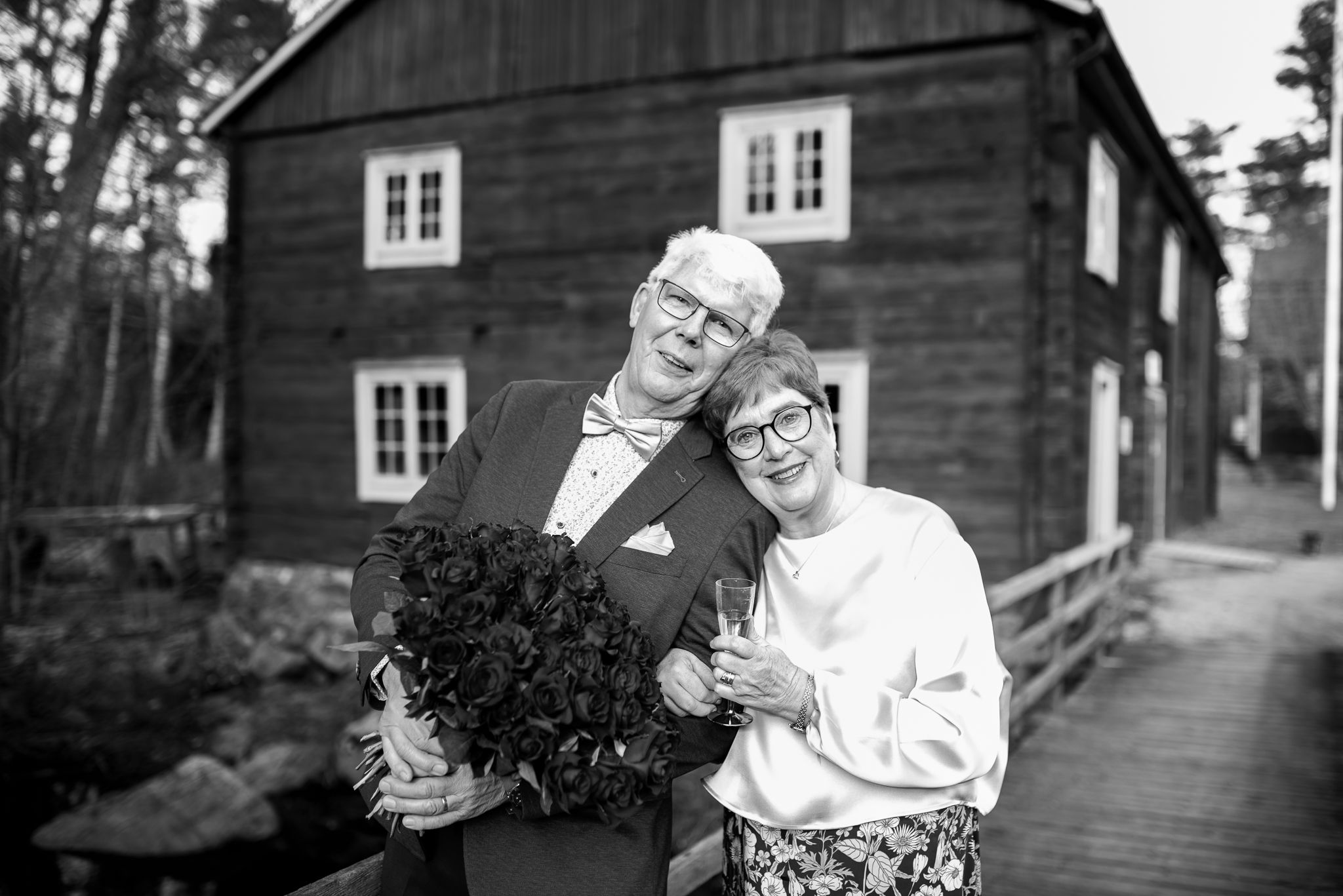 A couple poses with flowers in front of a rustic wooden building in black and white.