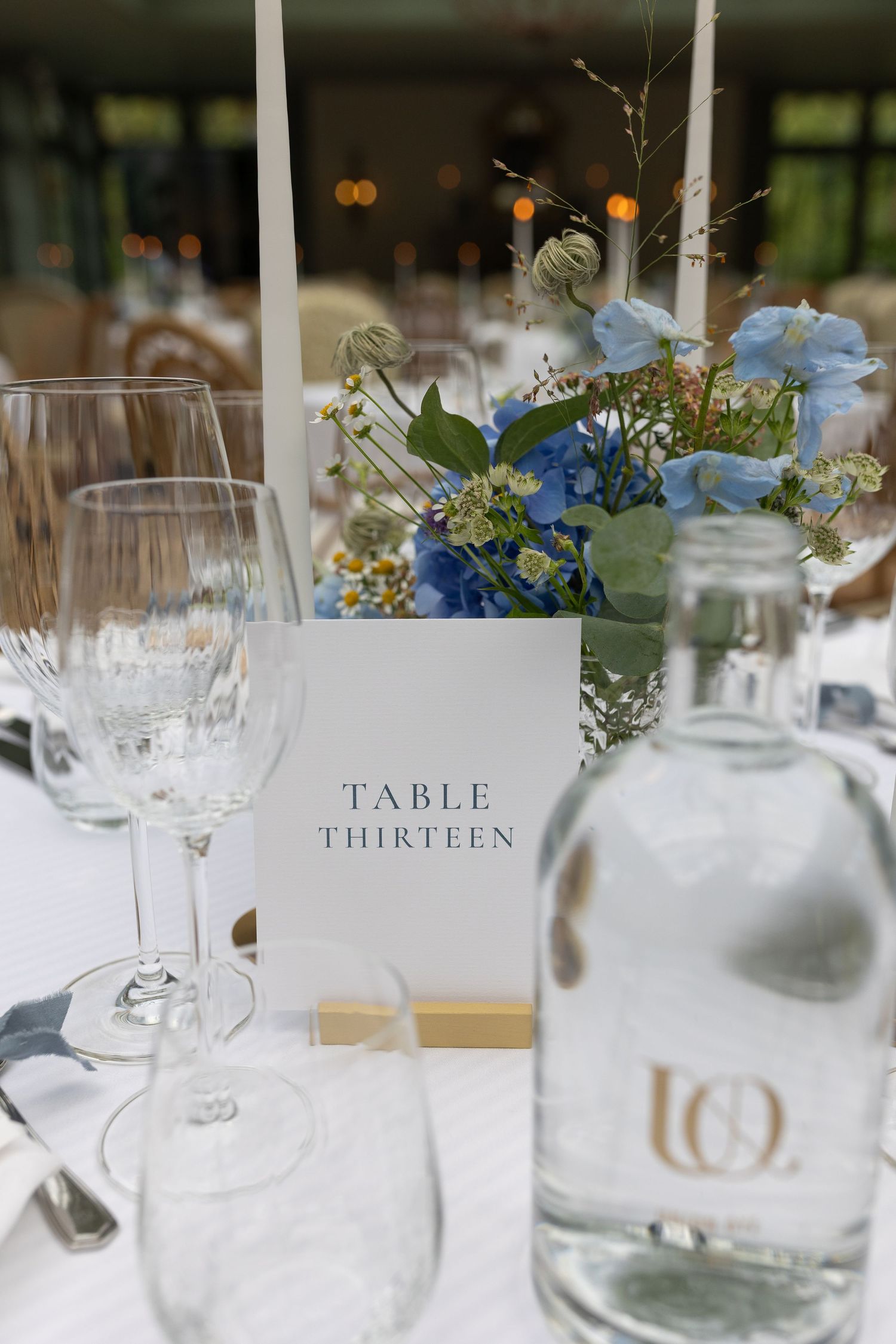 Elegant wedding table setting with 'Table Thirteen' card, wine glasses, and blue floral centerpiece.