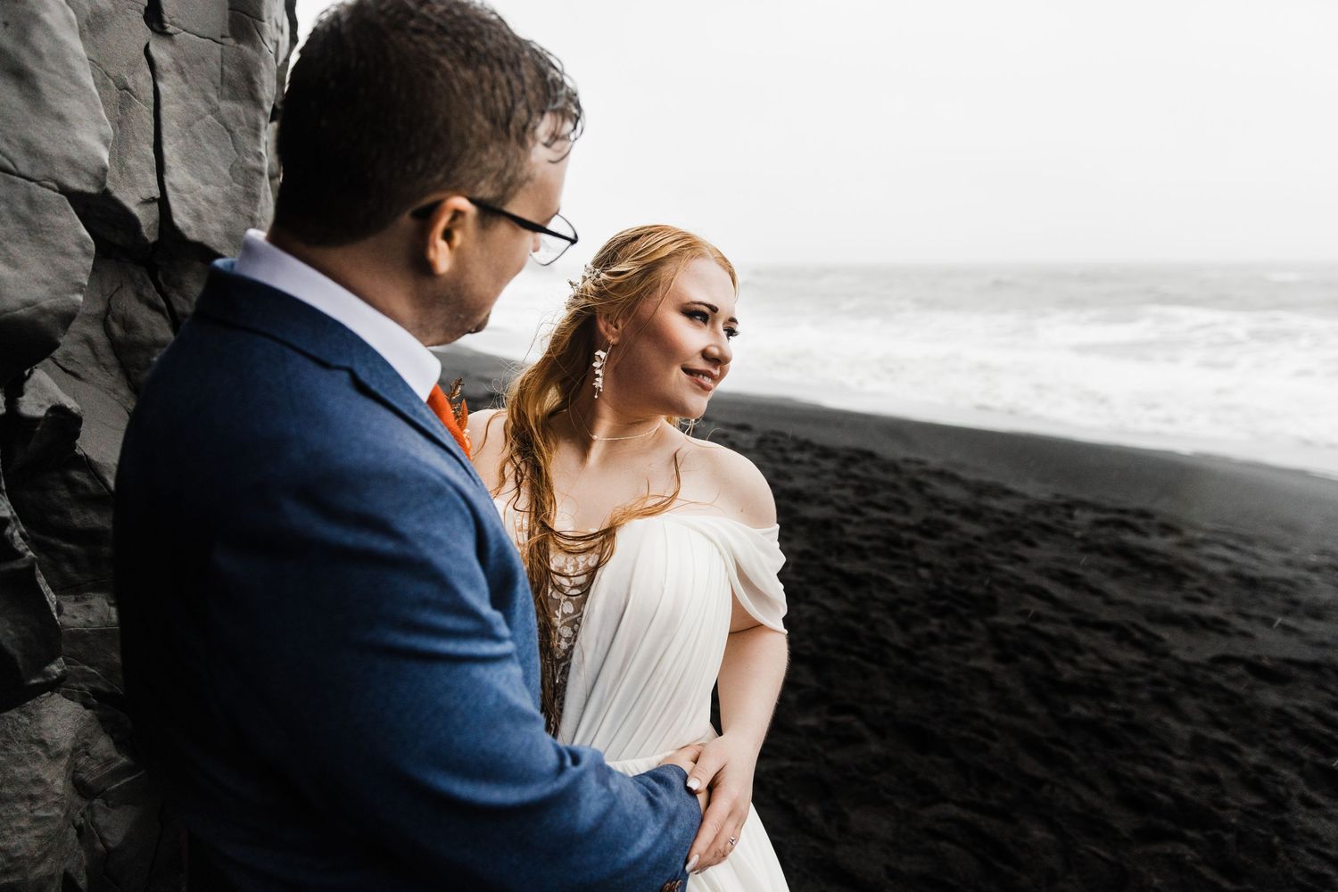 Couple stands against dramatic black sand beach with waves crashing in background.