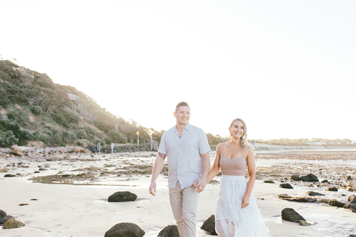A couple walks hand in hand along a scenic beach with cliffs and rocks in the background.