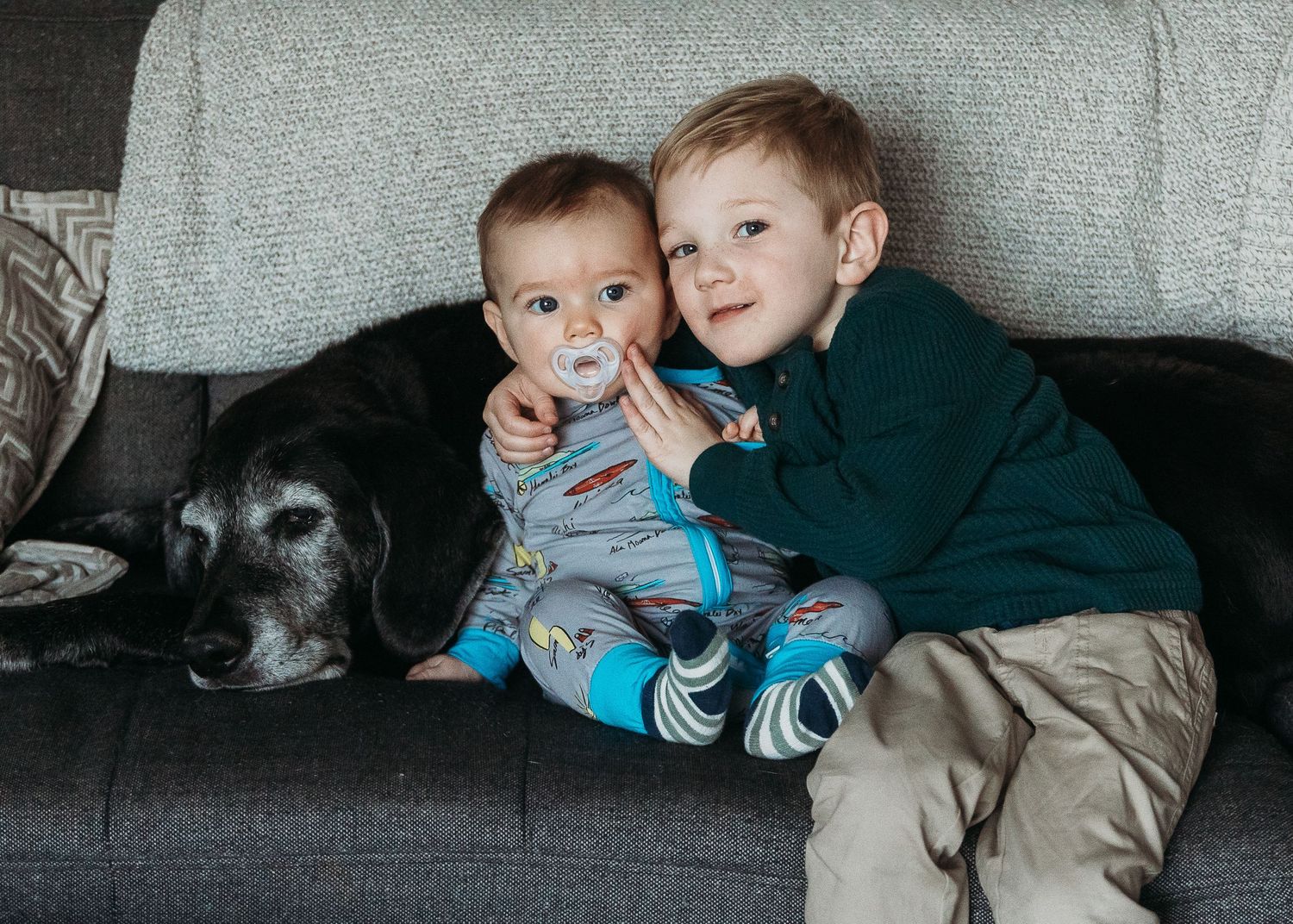 Two children cuddling on a grey couch with a black dog.