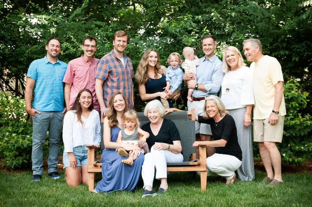 A large extended family poses together on an outdoor bench for a group portrait in a garden setting.