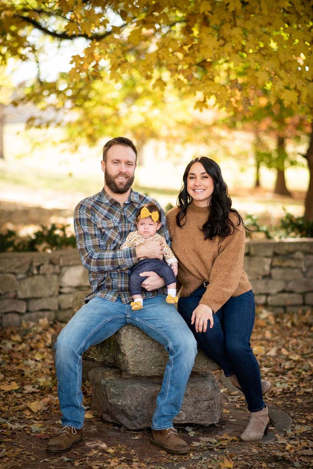 A small family poses together on a stone wall surrounded by autumn foliage and golden light.