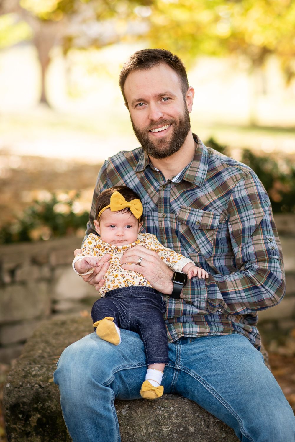 A baby wearing a yellow bow and floral top sits with a parent on a stone wall during a fall outdoor photoshoot.
