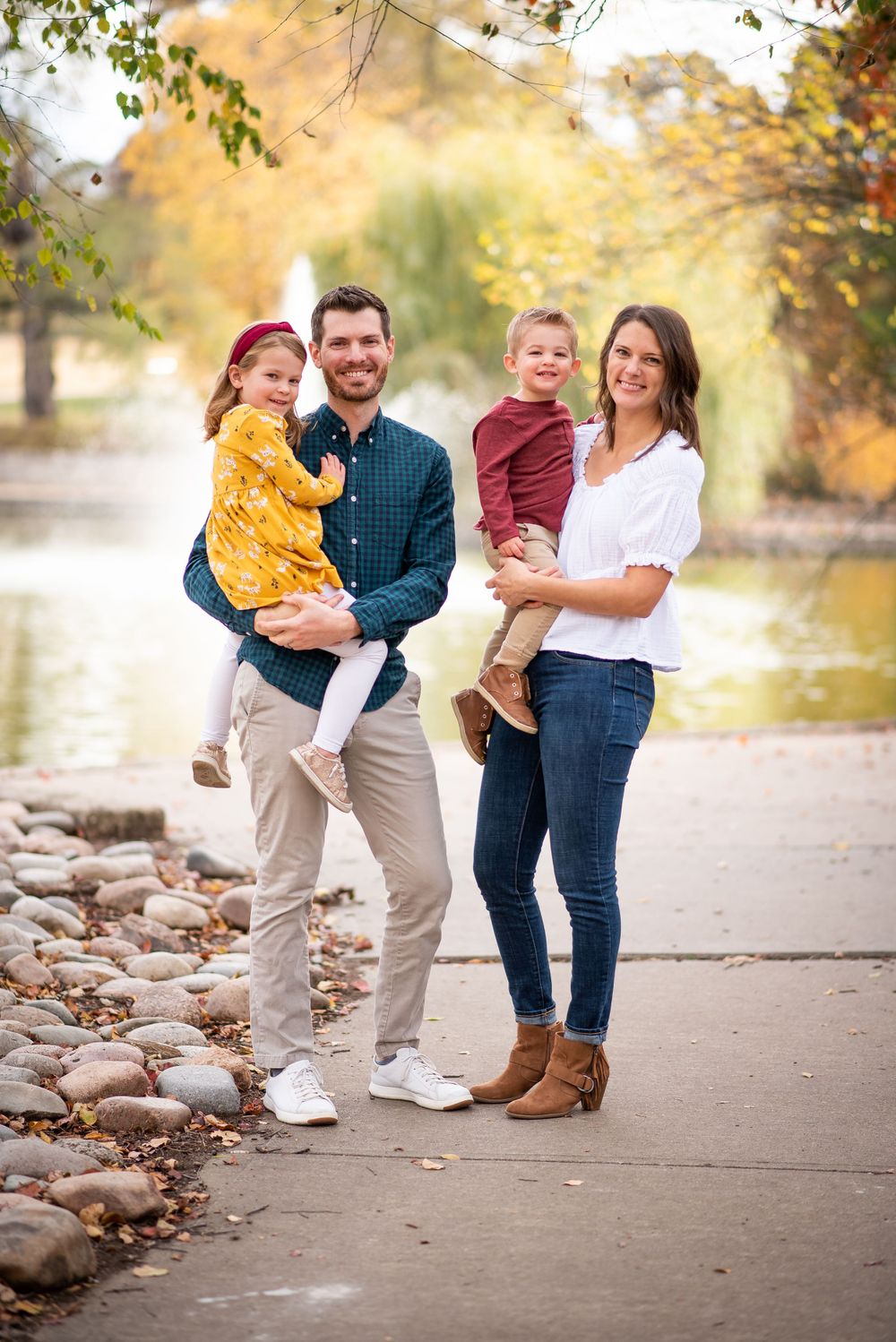 Family of four stands together on a path with autumn foliage and water in the background.