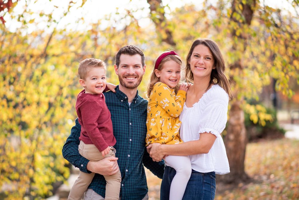 Family of four poses together in front of golden autumn trees during a fall photo session.
