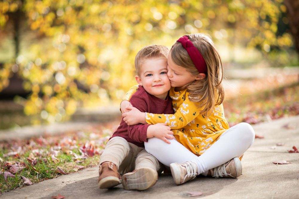 Two small children sit together on a path surrounded by fall foliage, sharing an affectionate moment.