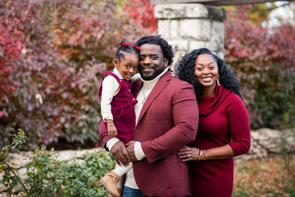 Family of three wearing burgundy outfits pose together against colorful fall foliage backdrop.