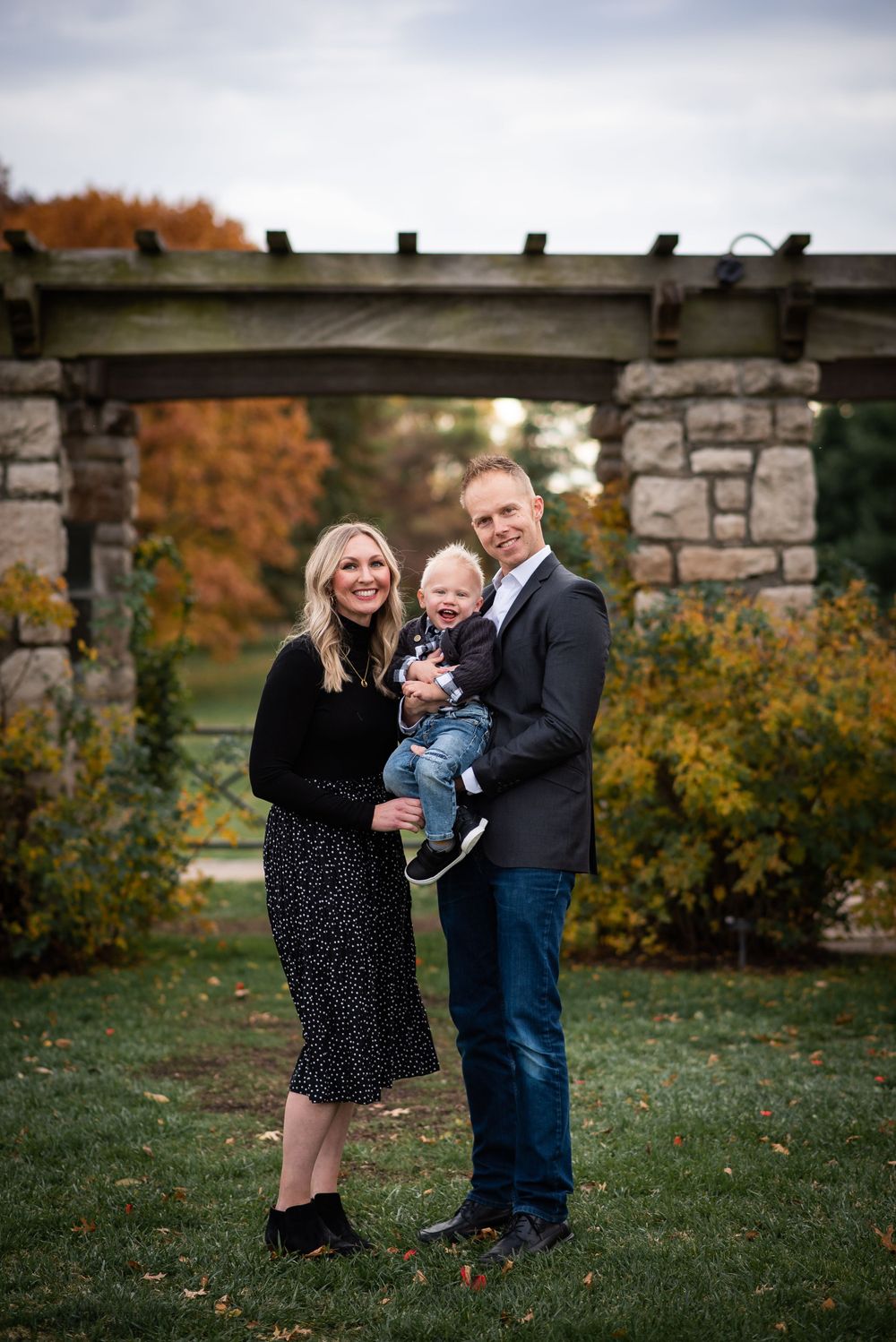 Family poses together under a stone archway during autumn, wearing coordinated dark outfits.