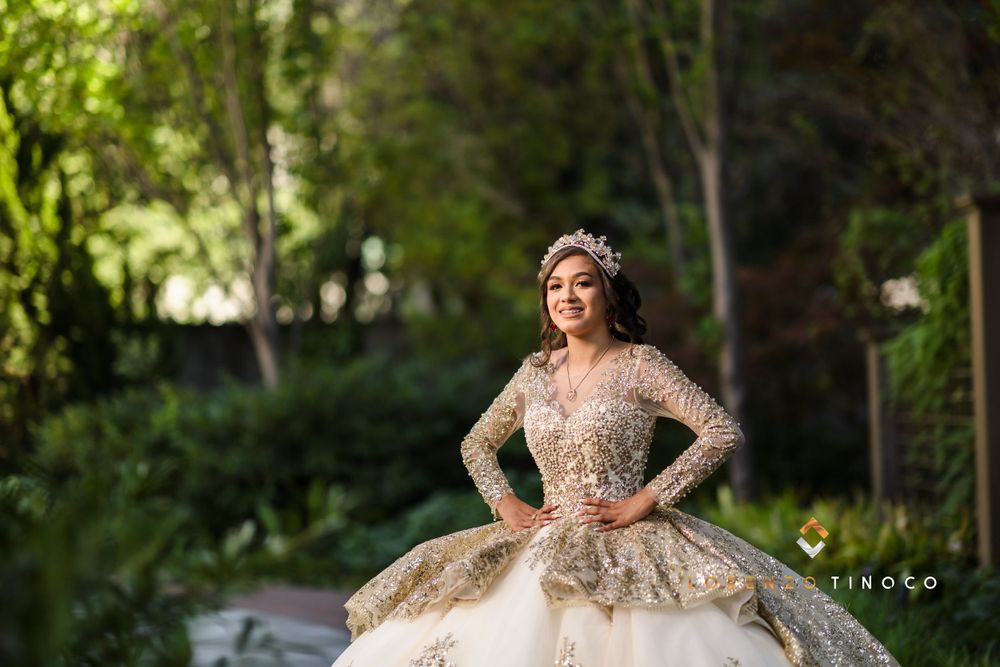 A model wearing a detailed gold and ivory ball gown poses in a lush garden setting.