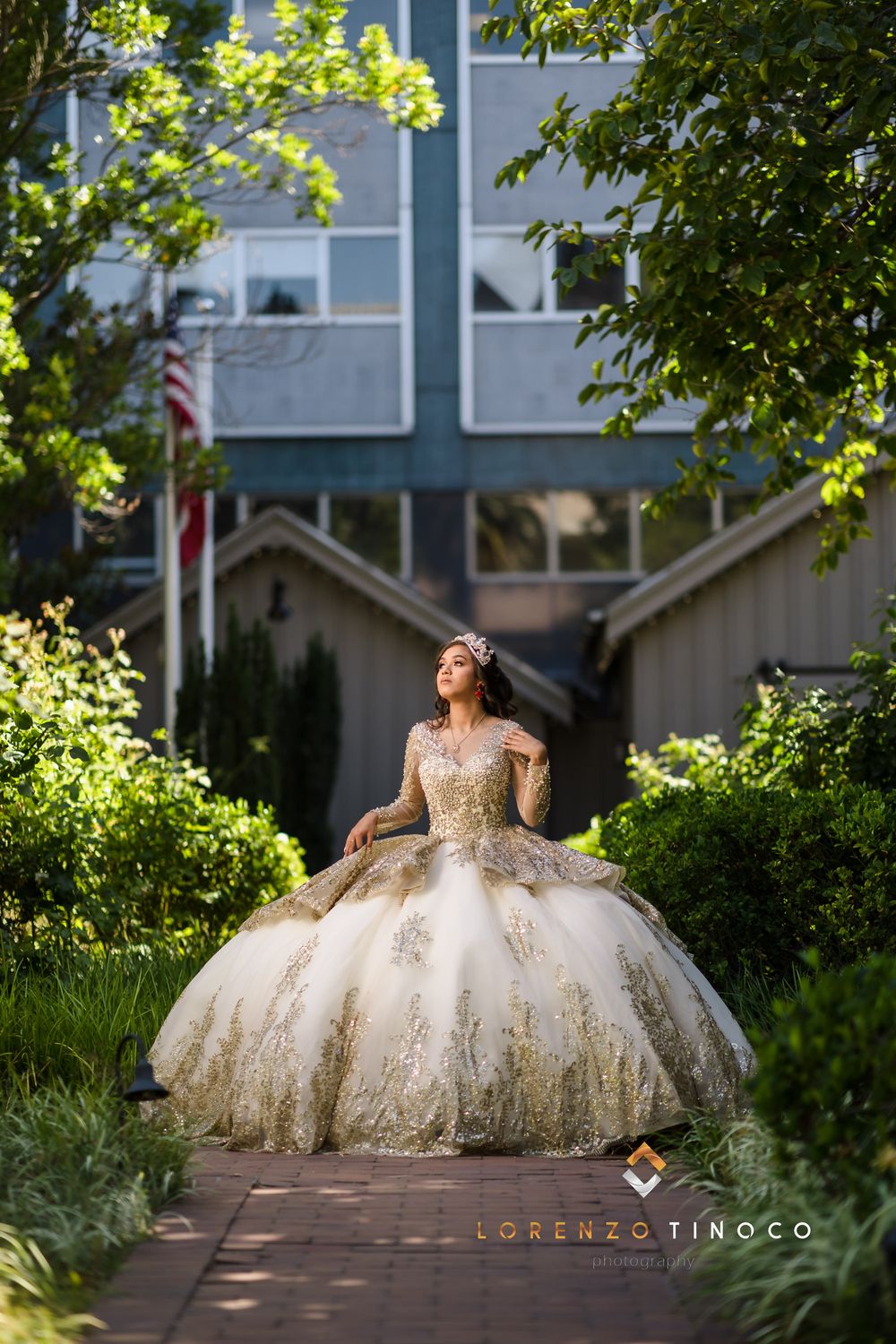 A model in a gold and ivory ball gown poses dramatically in dappled sunlight on a garden path.