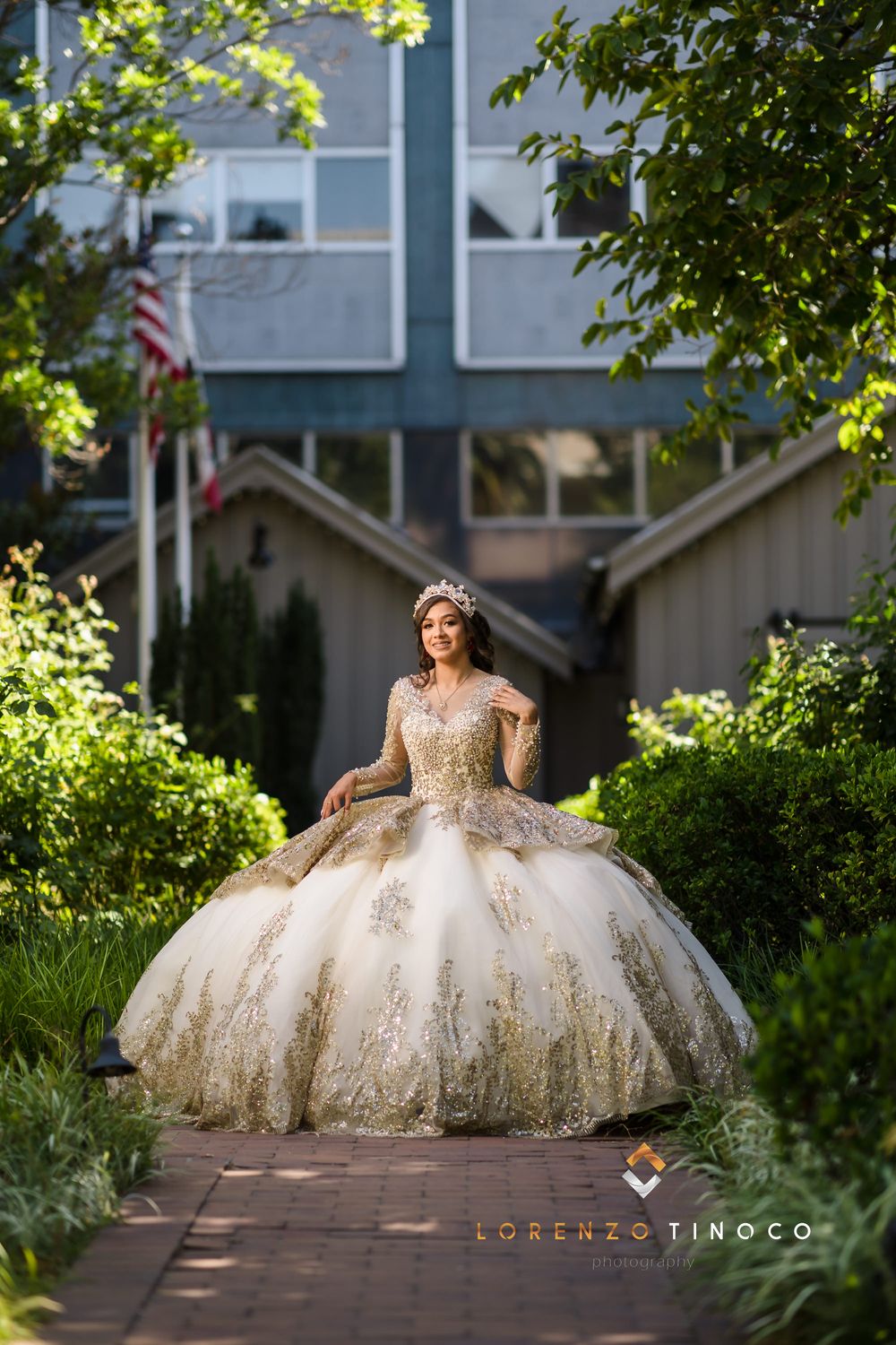 A model in an embellished ivory ball gown stands on a garden path with a building backdrop.