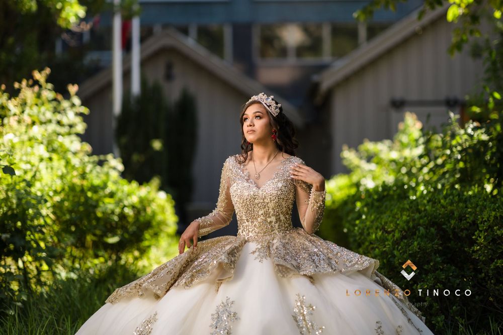 A model in an ornate gold ball gown poses regally in a sunlit garden setting.