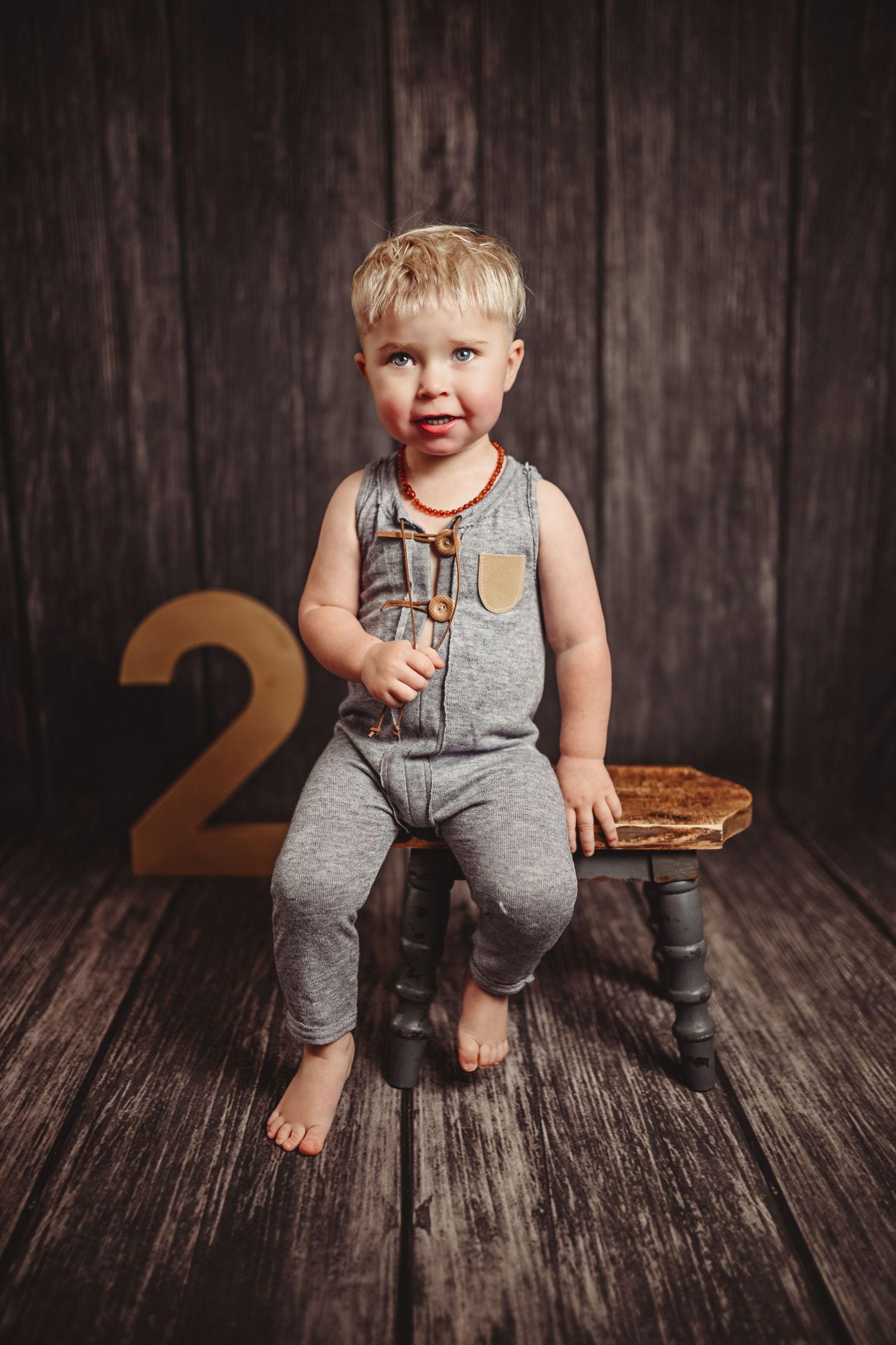 Toddler in grey overalls sitting on dark wooden floor with number 2 prop.