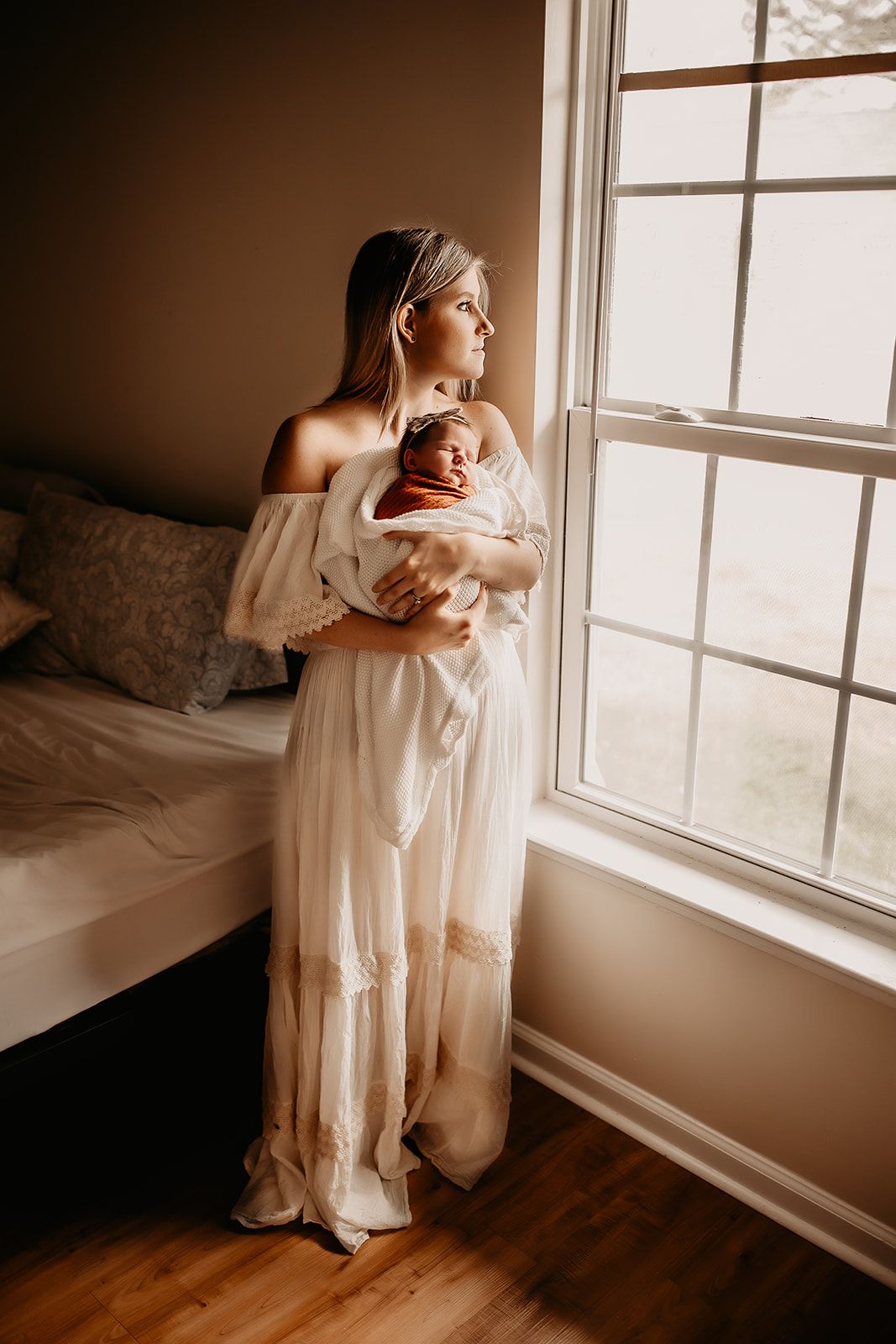 A mother stands by her bedroom window holding her newborn baby girl during their in home newborn photoshoot.
