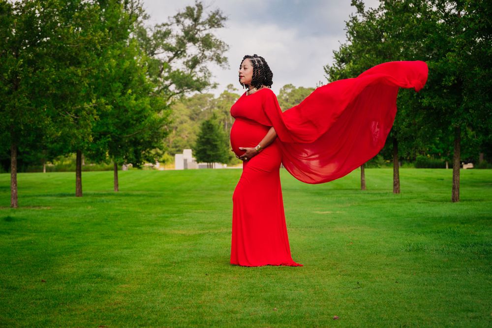 A flowing red dress billows dramatically in the wind as someone poses gracefully in a green park setting.
