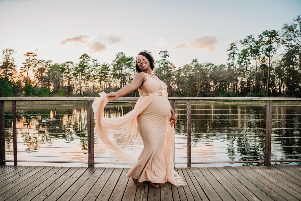 A graceful figure in a flowing beige gown poses on a wooden dock at sunset beside a tranquil lake with pine trees.