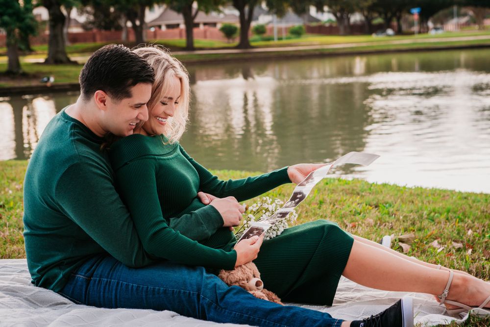 A couple in matching green sweaters sits on a blanket by a pond reading together on a peaceful afternoon.