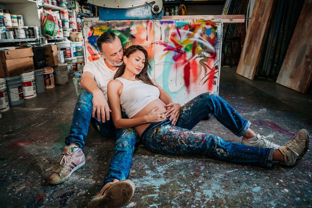 Two people pose casually on paint-splattered floor with colorful graffiti wall background in urban studio setting.