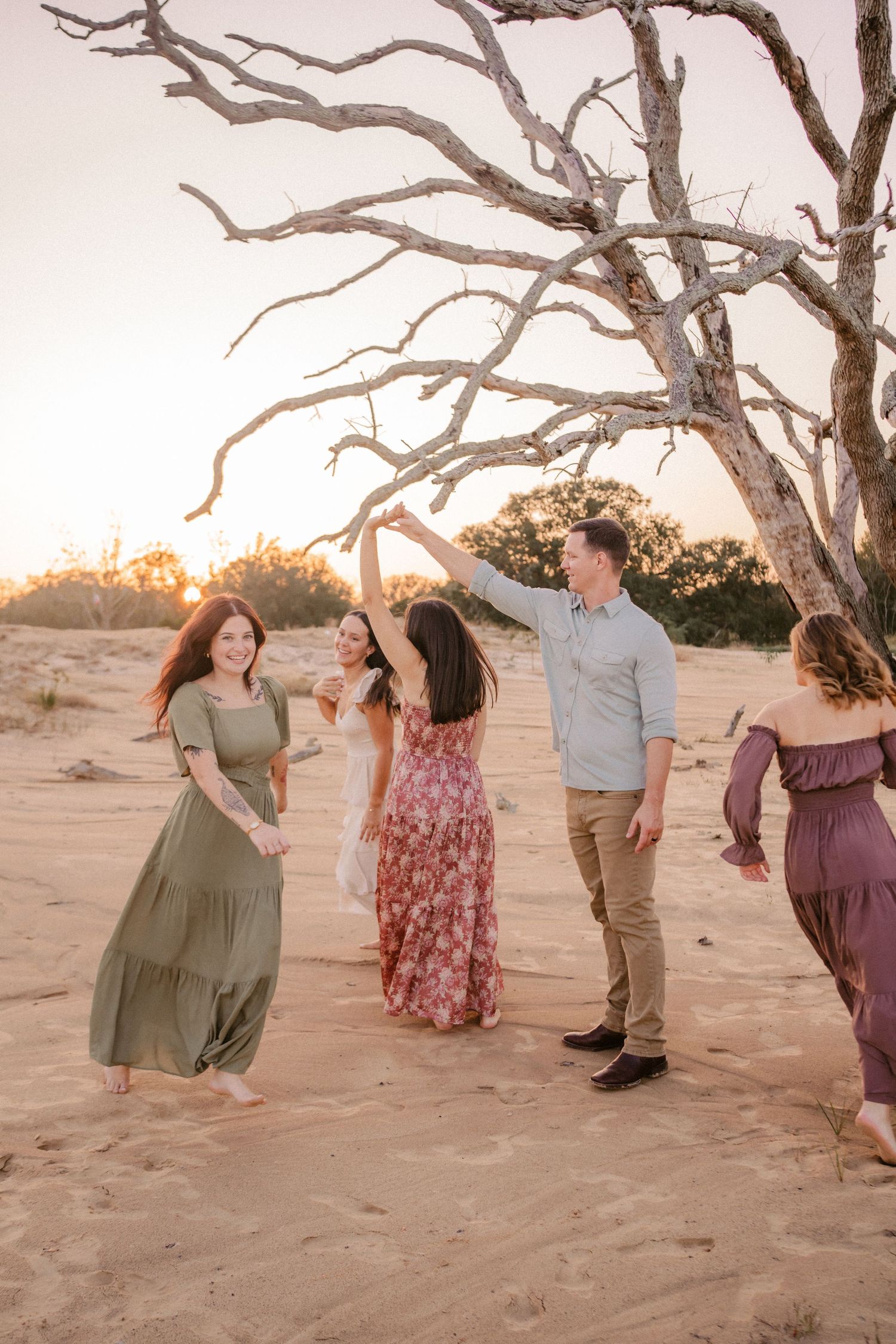 People in flowing dresses gather around a bare tree at sunset in a desert landscape.