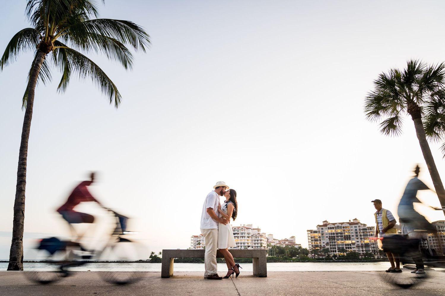 Romantic beachside scene with palm trees and cyclists passing by at sunset.