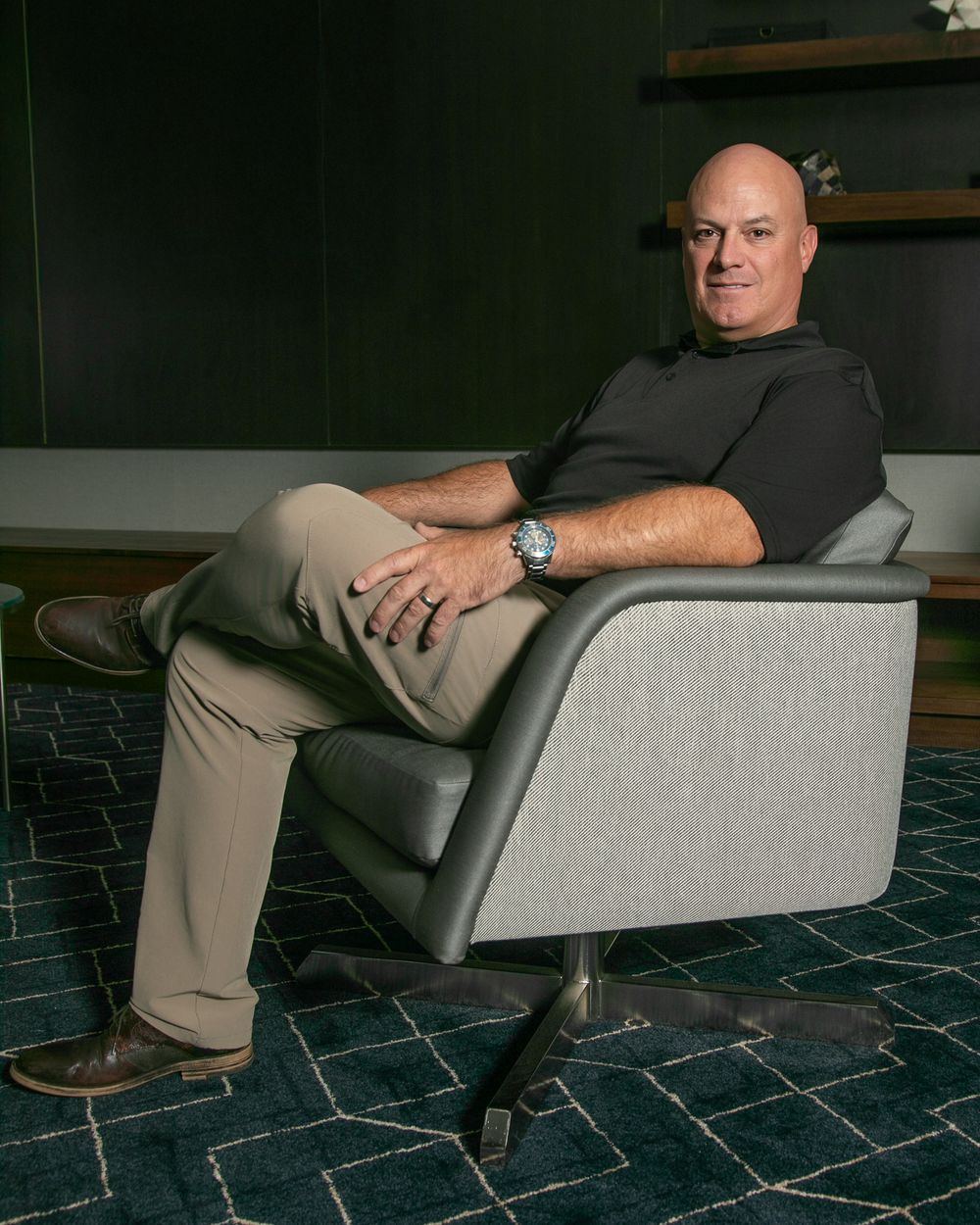 A person sits in a gray upholstered armchair on a geometric patterned floor in a dark room.