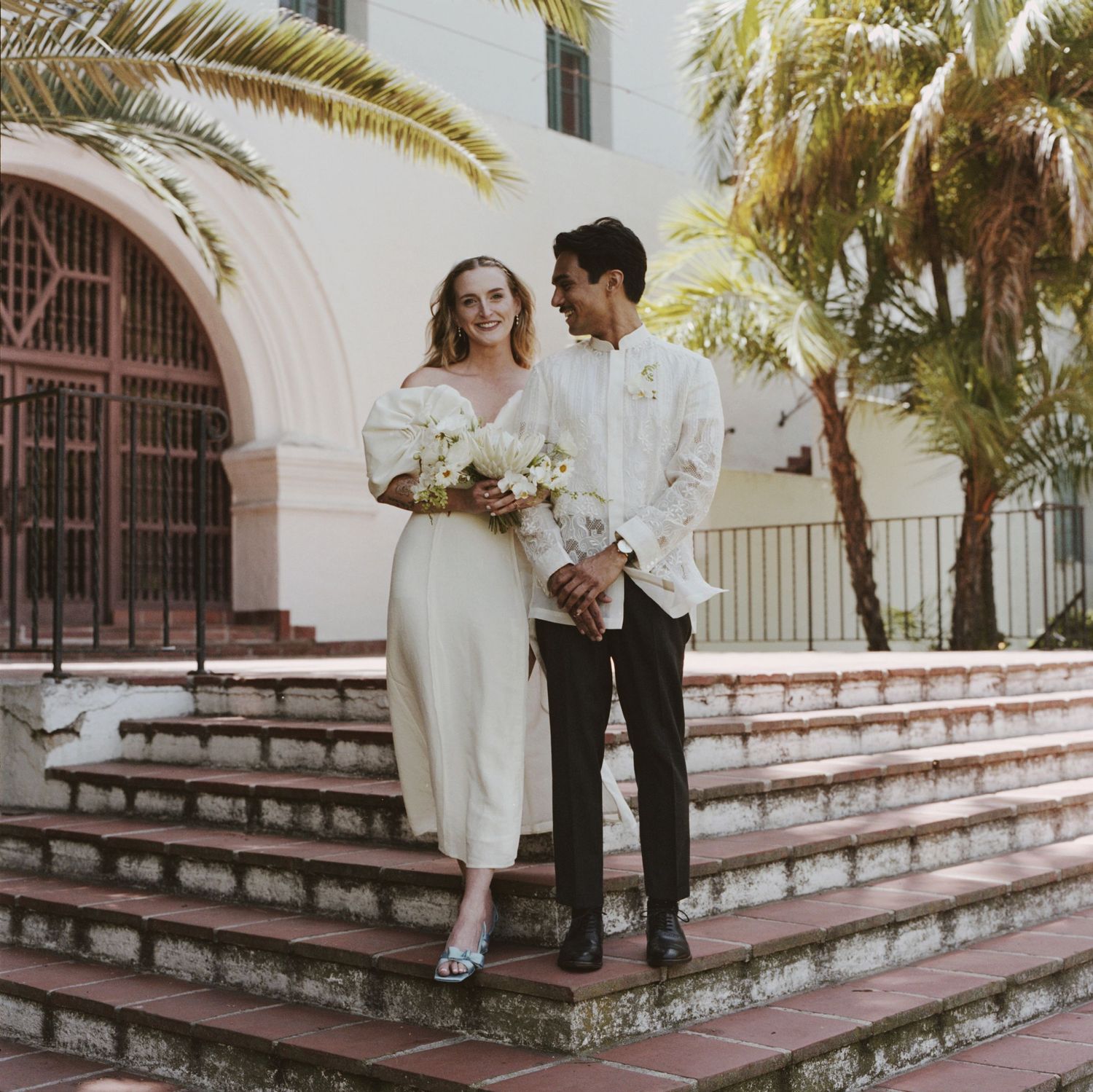 Newlyweds stand on Spanish colonial style church steps surrounded by palm trees.
