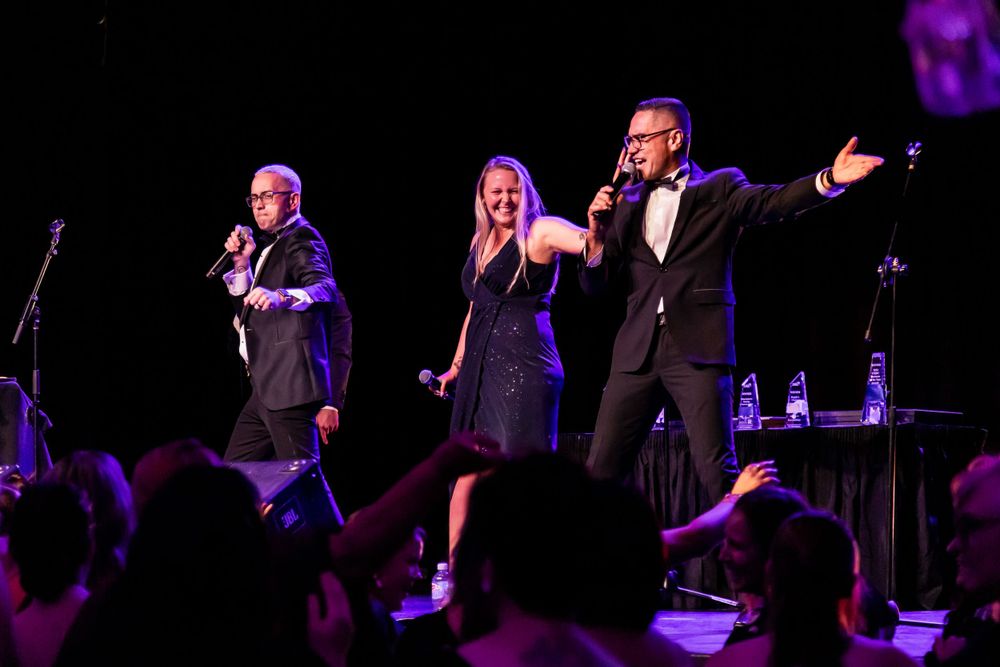 Performers singing and gesturing dramatically on a darkened stage with purple lighting.