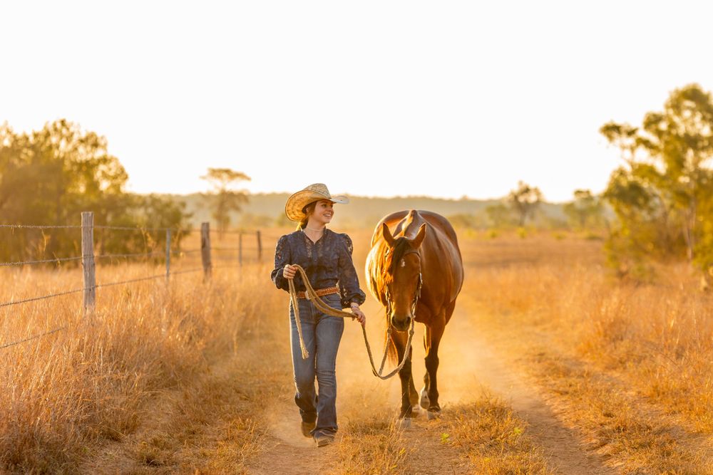 Person walks with horse along dusty country path during golden sunset.