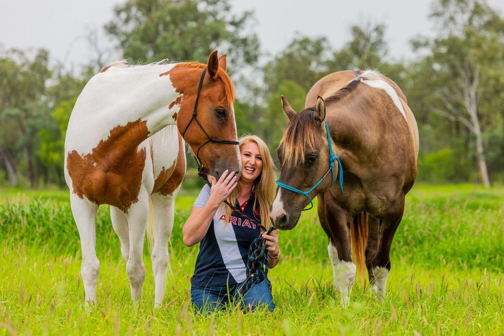 A person in overalls poses with two horses, one paint and one brown, in a lush green field.