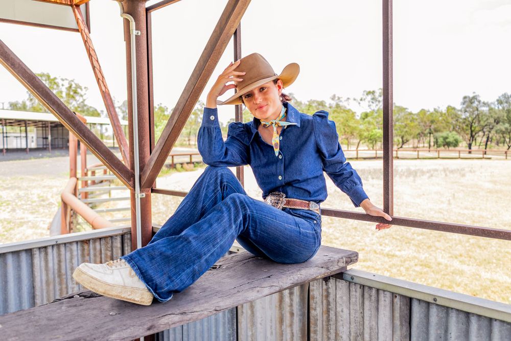 A person in western attire and cowboy hat sits casually on a wooden railing at a ranch.