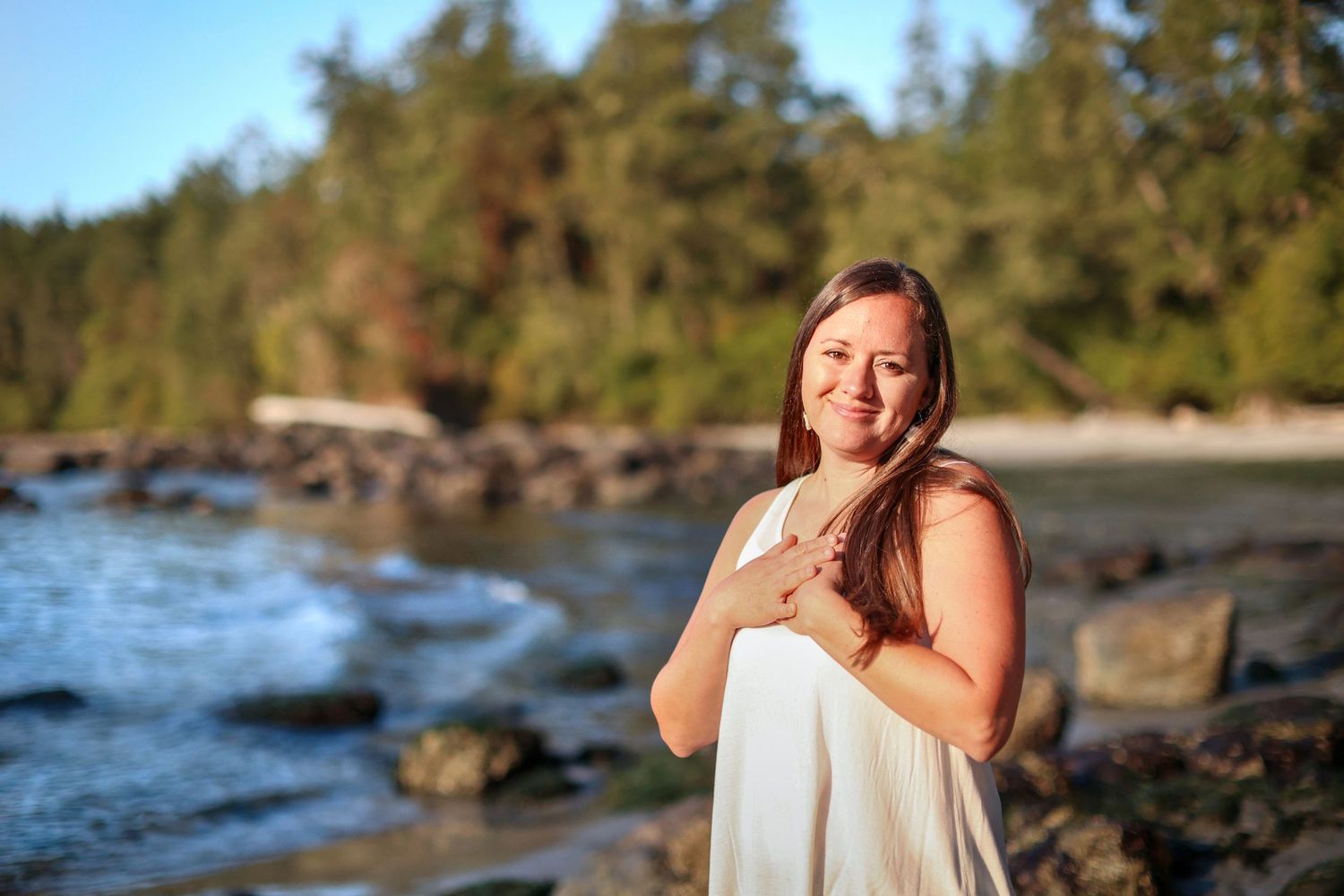 Smiling woman in white dress on rocky beach with forest backdrop at sunset.