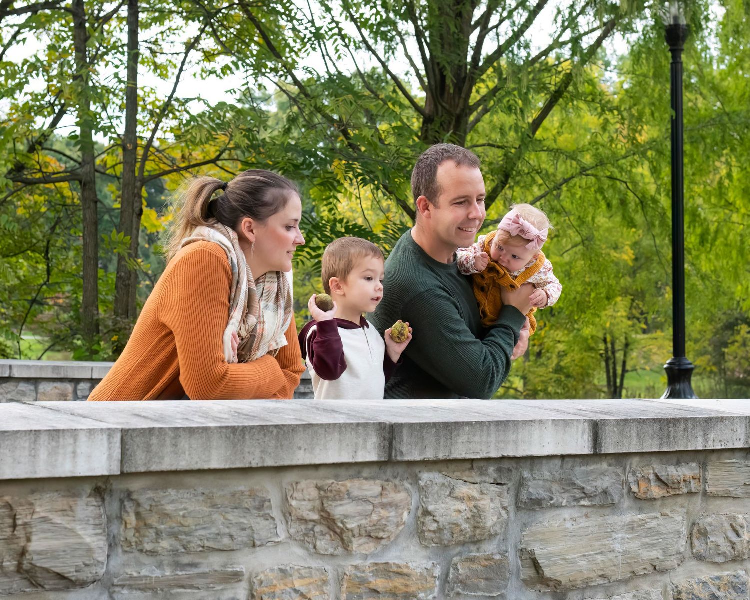 A family of four leans together against a stone bridge with green trees in the background.