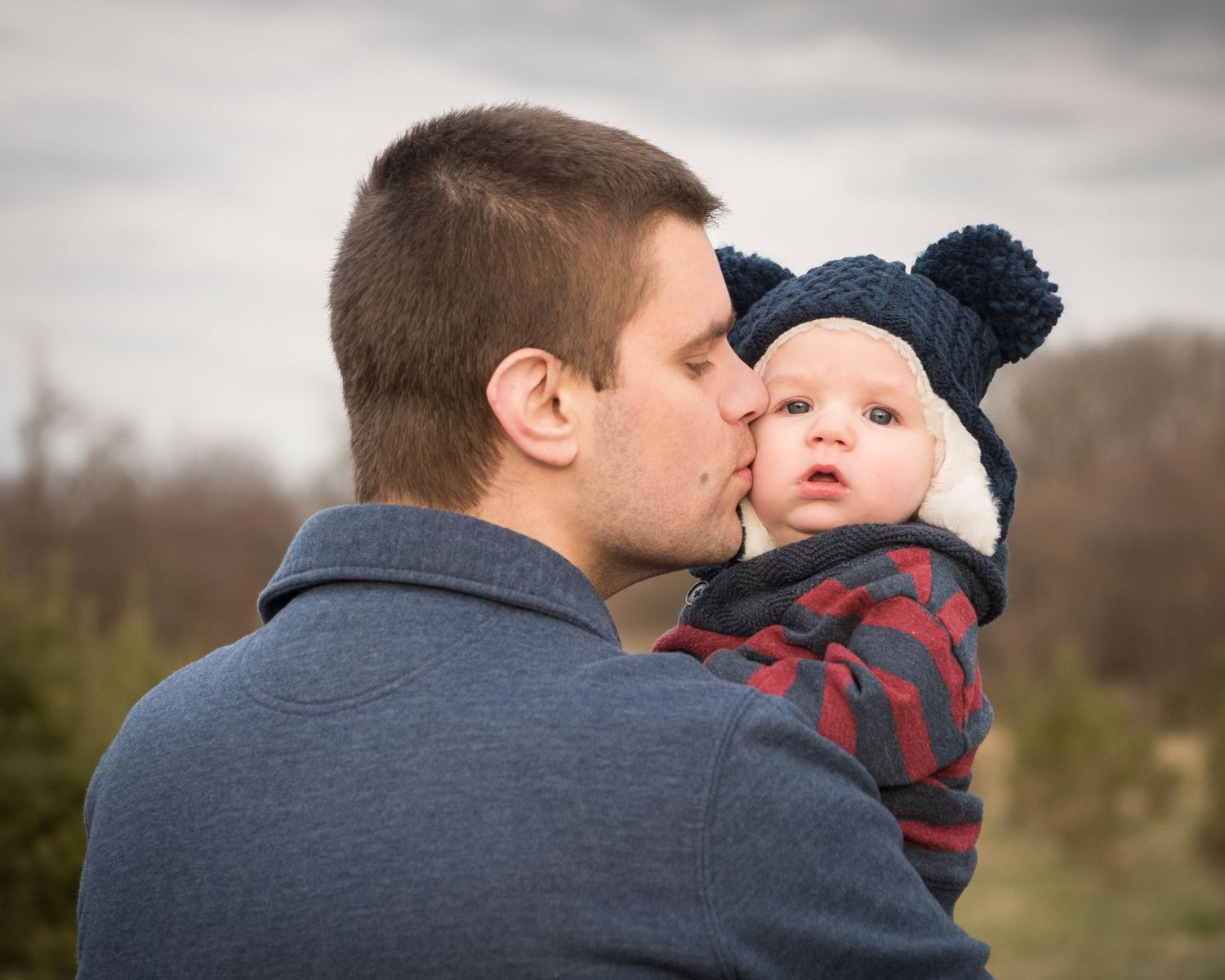 A father in a gray sweater kisses his baby who is wearing a blue knit hat and plaid shirt against a blurred winter backdrop.
