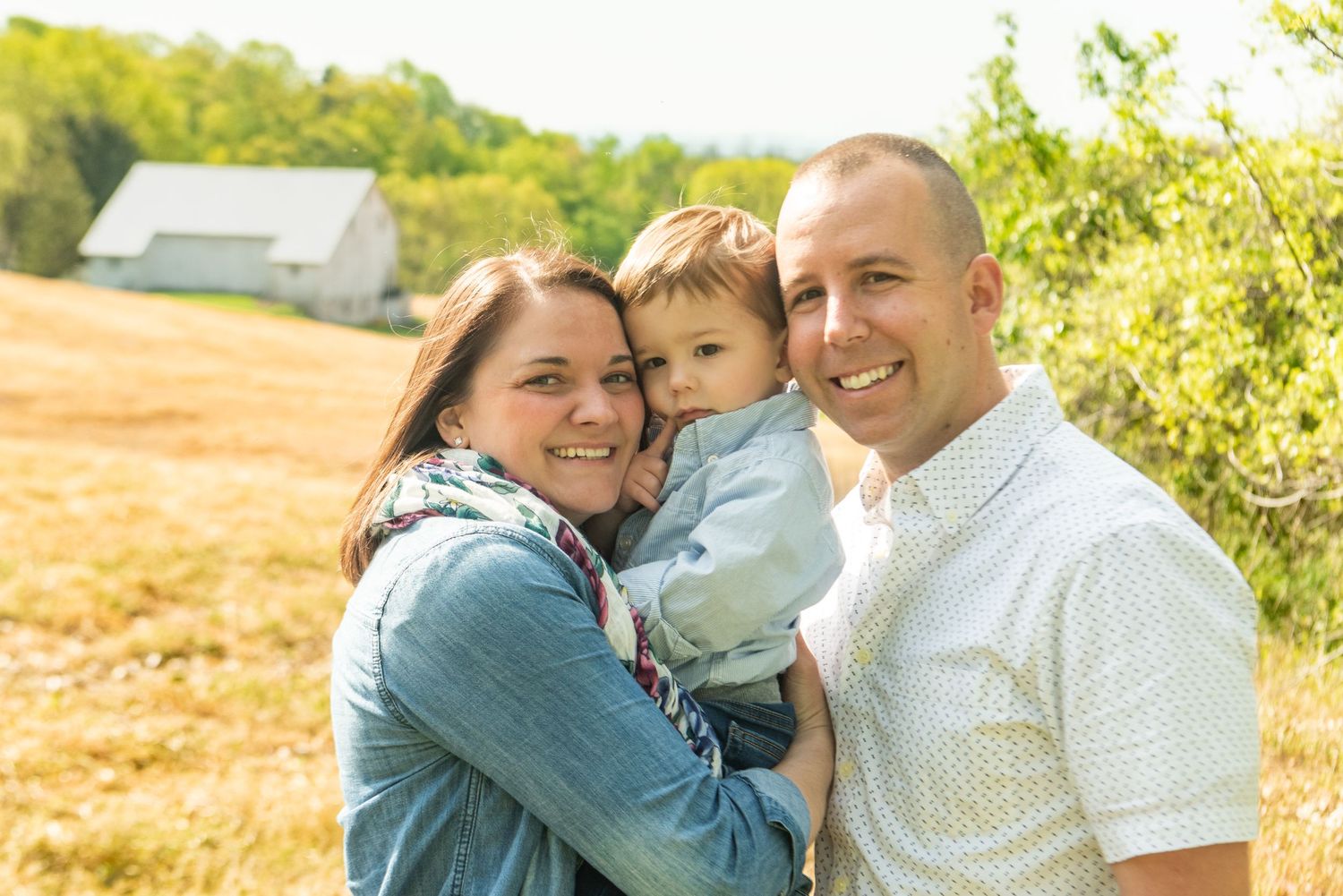 Three people embrace and smile together outdoors on a sunny day with a golden field and barn in the background.