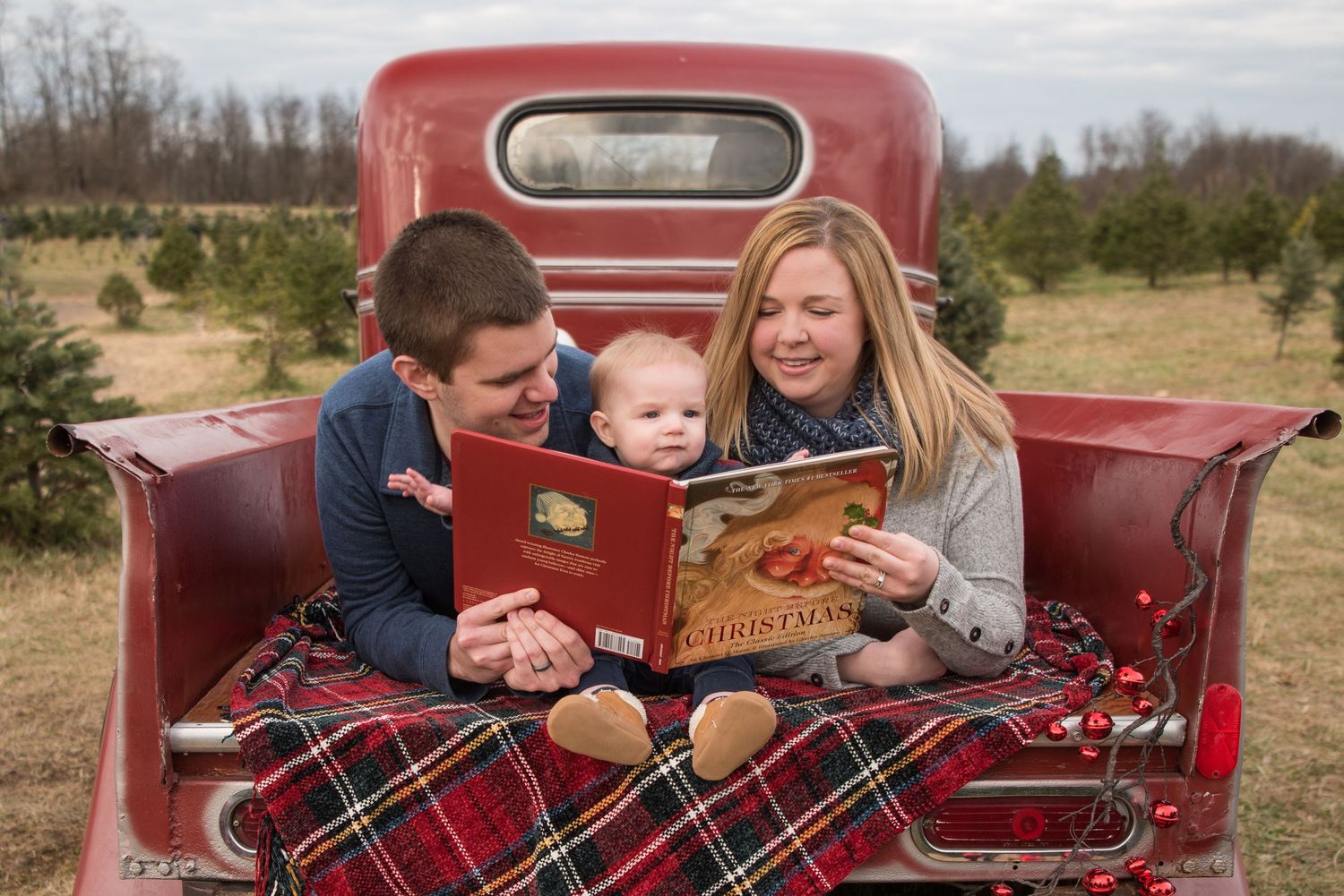 Family reads a book together while sitting in the back of a vintage red pickup truck with a plaid blanket.