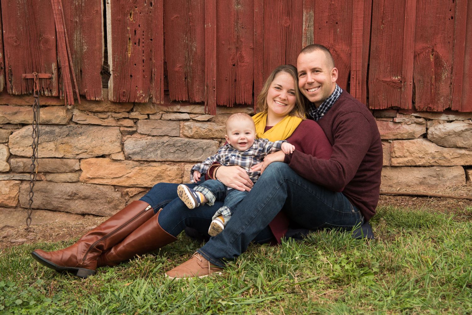 A family sits together on grass against a rustic red barn wall with stone foundation on an autumn day.